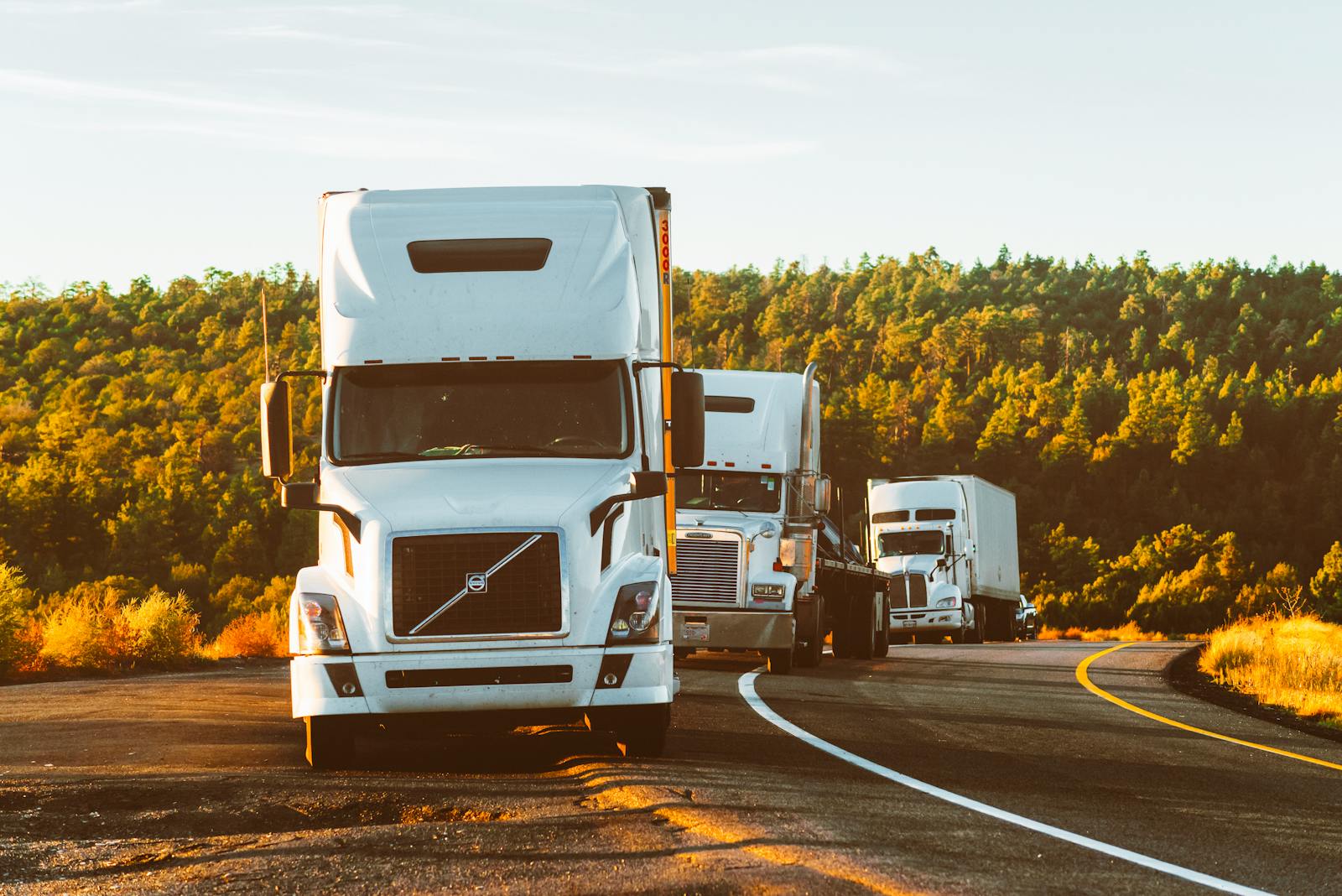 Three semi trucks driving on a highway through a forested landscape, commercial truck